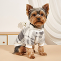 A small fluffy brown dog wearing a soft fuzzy sweater with a gray, white, and beige plaid pattern and white ribbed trim, posed indoors against a light neutral background.